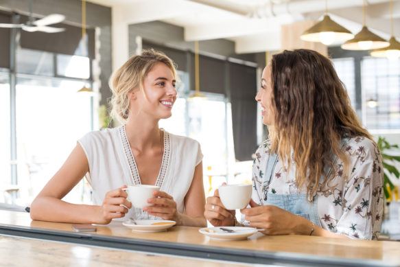 Two beautiful young women holding cup of coffee and talking to e
